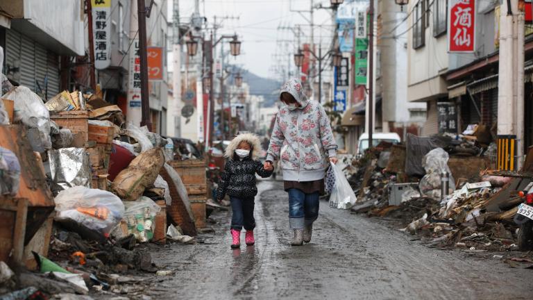 An adult and child walk hand in hand along a street in an urban area on a path cleared through debris. 