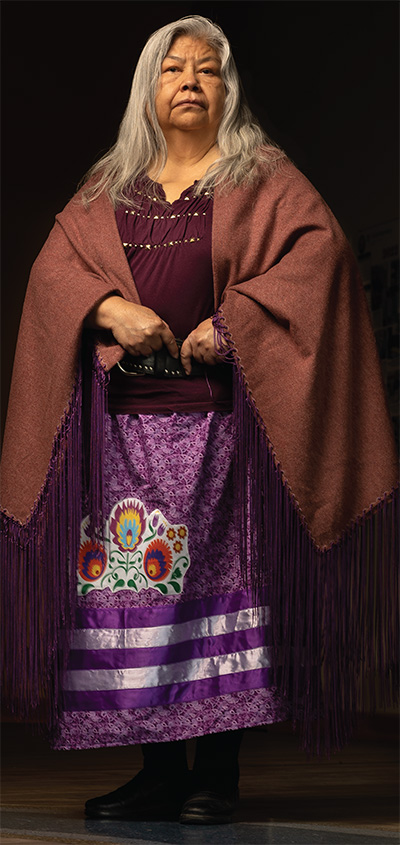 woman with long grey hair stands facing camera, wearing a shawl draped across her shoulders and a long purple embroidered skirt