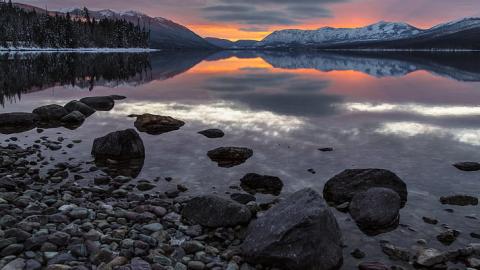 A sunset is reflected in still water with rocks in the foreground and snowy mountains in the distance. 