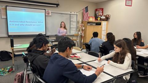 A young woman stands at the front of a high school class in front of a screen with a slide reading "Antimicrobial Resistance: What is it and why should we care?"
